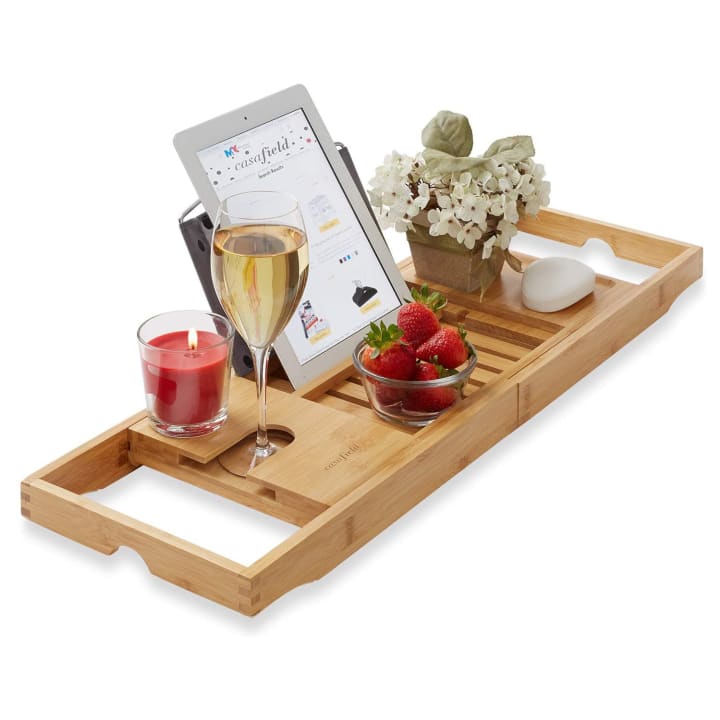 Casafield Bamboo Bathtub Caddy with a candle, wine glass, flowers, fruit, and a tablet on a white background