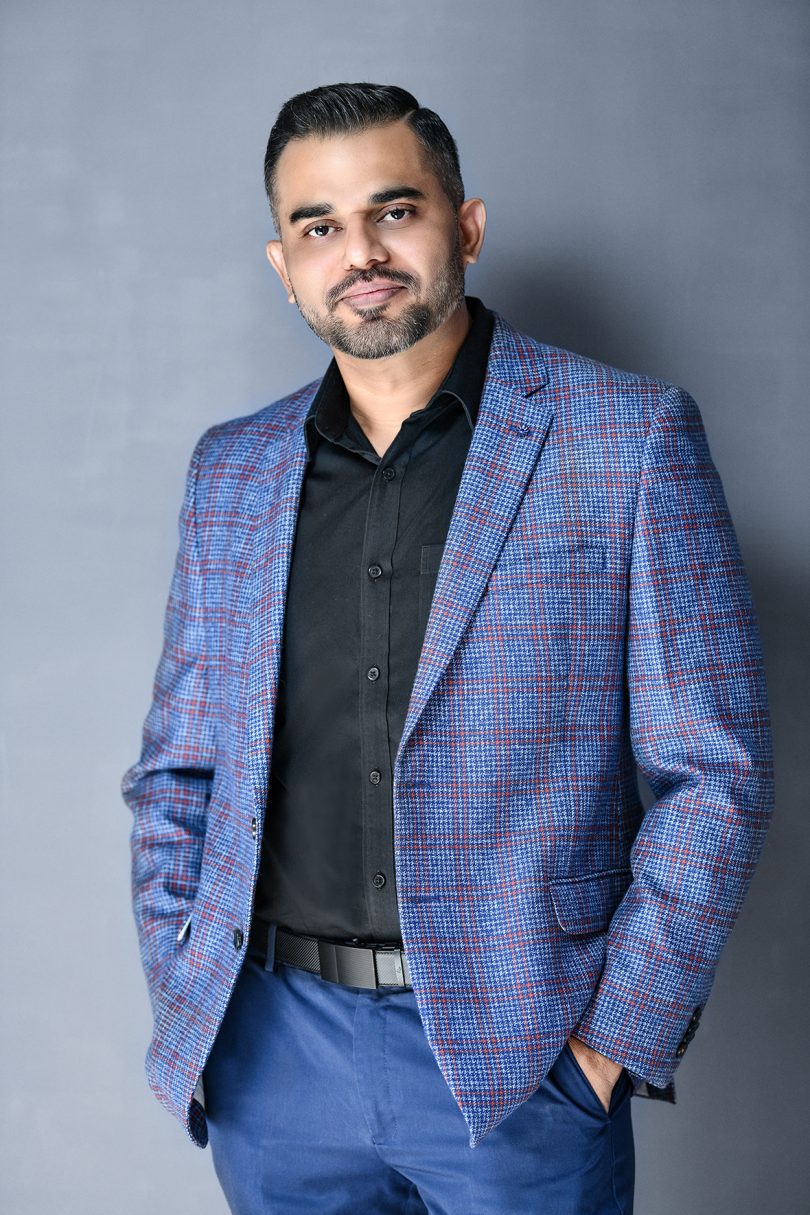 brown-skinned man with salt and pepper hair wearing a black button up shirt and blue plaid blazer while looking at the camera