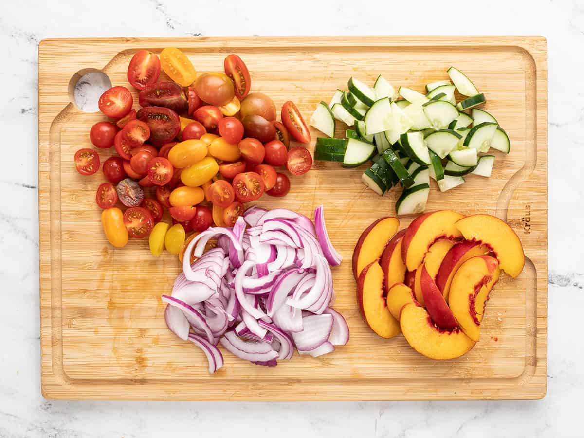 Overhead shot of chopped onions, tomatoes, cucumbers, and nectarines on a wood cutting board.